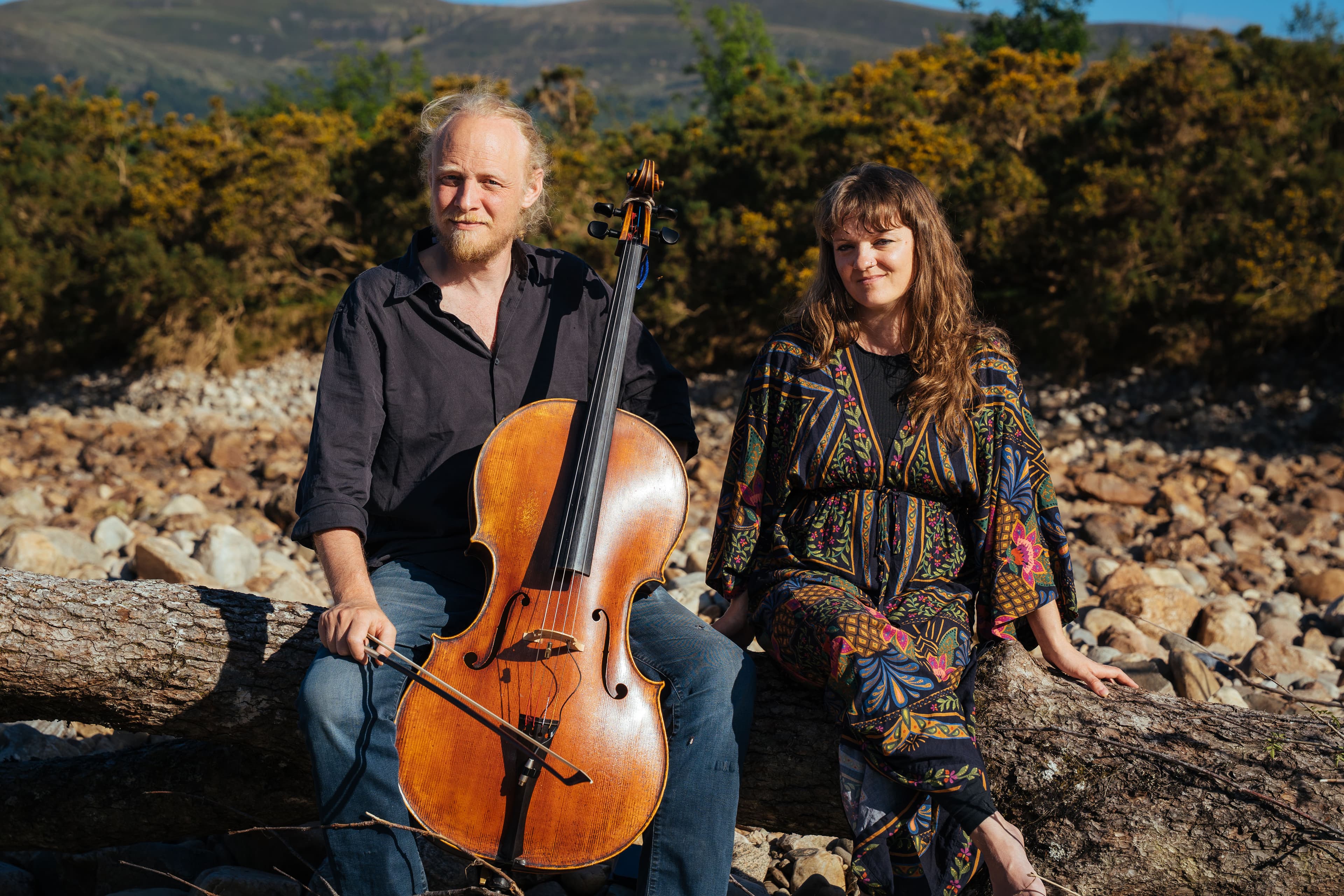 A man and a woman sit on a log in the sunshine, the man is blonde and bearded and holds a chello, the woman has long brown hair and wears a floral dress. They sit side by side lookingat the camera.